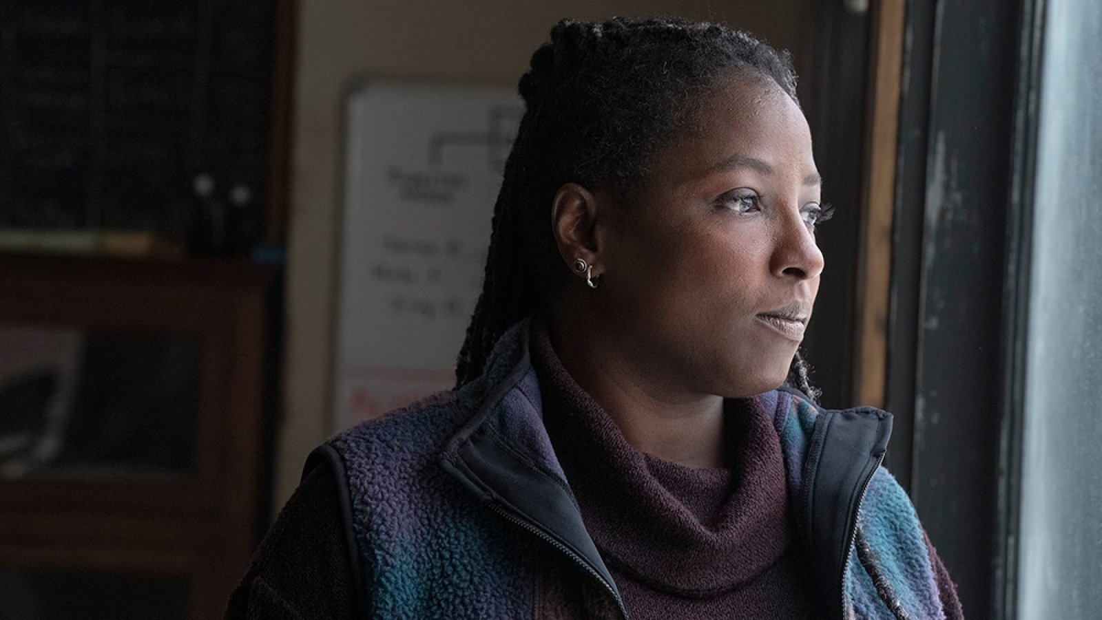 A woman stands in a room staring out the window, looking thoughtful.