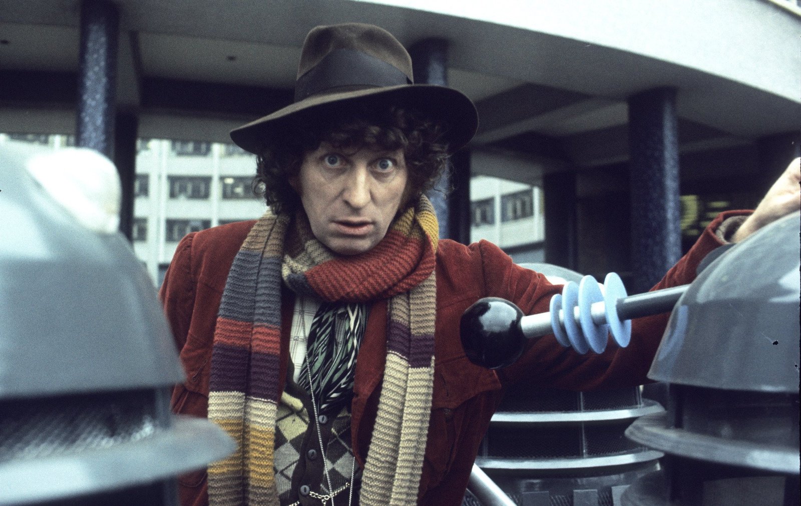 Tom Baker in hat and scarf looks directly at the camera between Daleks